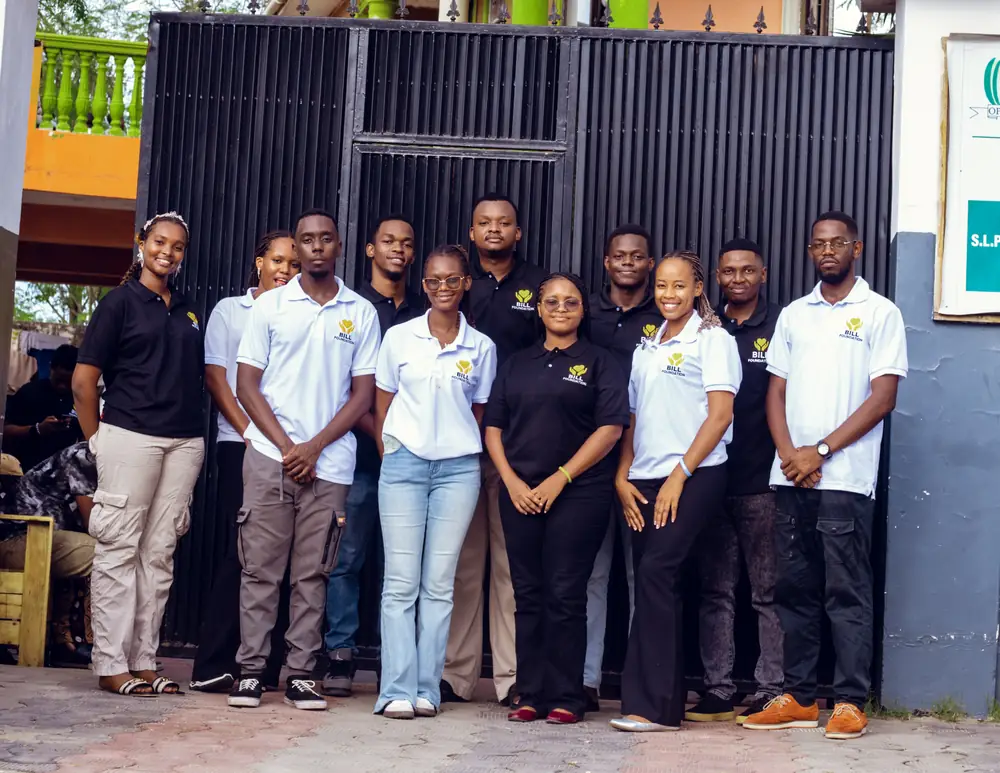 Members of the Bill Foundation TZ leadership team in branded black and white polo shirts, standing in front of a black gate